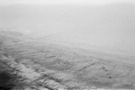 Salt Coast, 2019. Black-and-white analogue photograph showing a vast, pale coastal landscape with subtle ground textures, scattered dark traces and a nearly disappearing horizon.