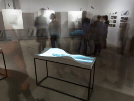 Long-exposure view of a sand simulation projected onto the sandhouse sculptural model, surrounded by blurred exhibition visitors