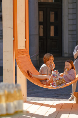 Two children seated together on an orange suspended seating element within Dome Next Door, 2024, an urban art installation in Riga Old Town, Latvia.