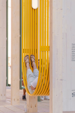 Person reclining in a yellow suspended seating element within Dome Next Door, 2024, a timber urban art installation in Riga Old Town, Latvia.