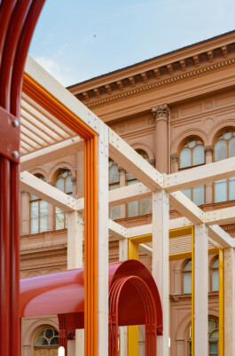 Detail of Dome Next Door, 2024, showing the exposed timber structure with red, orange and yellow architectural elements against the historic façade of Riga Old Town, Latvia.