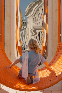 Child interacting with an orange woven seating element and distorted reflective surface within Dome Next Door, 2024, an urban art installation in Riga Old Town, Latvia.