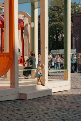 Visitors interacting with Dome Next Door, 2024, an urban art installation in Riga Old Town, Latvia, with green seating elements integrated into the timber structure.