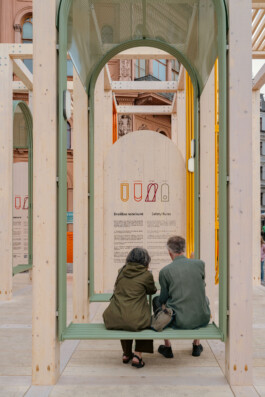 Two people seated on a green bench within Dome Next Door, 2024, framed by an arched green element with the installation’s safety rules visible behind them in Riga Old Town, Latvia.