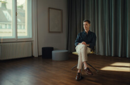 Portrait of Jurgis Gečys seated in a softly lit interior in Vienna, with one of his artworks displayed on the wall behind him. Natural daylight enters through a large window, creating a quiet architectural atmosphere.